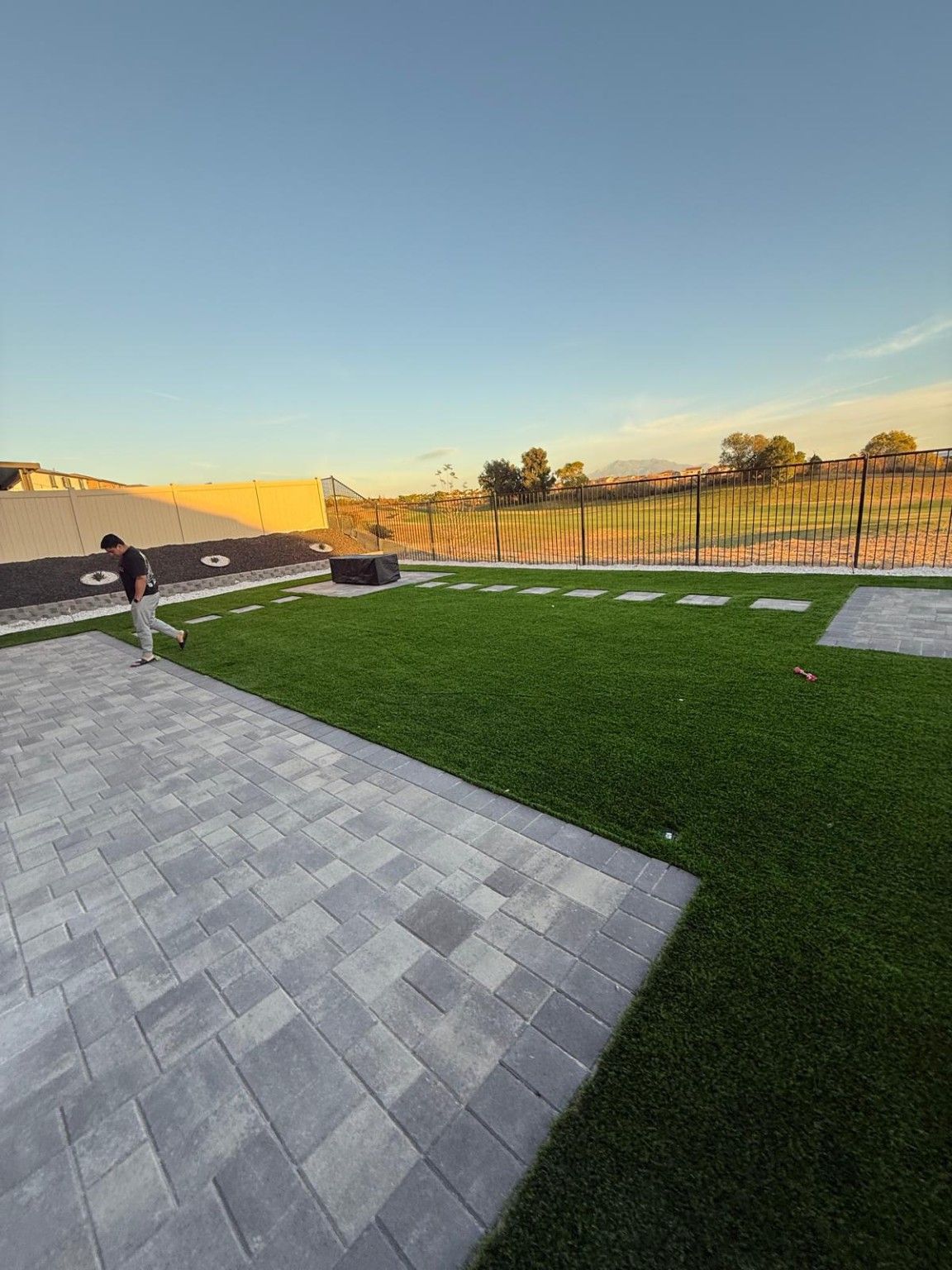 Gray paver patio beside artificial turf, with stepping stones along the rear fence and a raised planter at left