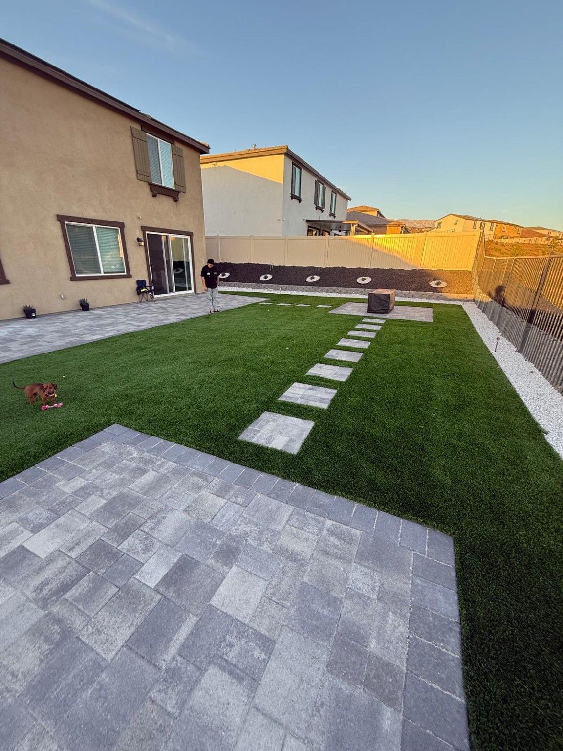 Rectangular stepping stones cross artificial turf toward a small paved area, with a raised rock planter behind