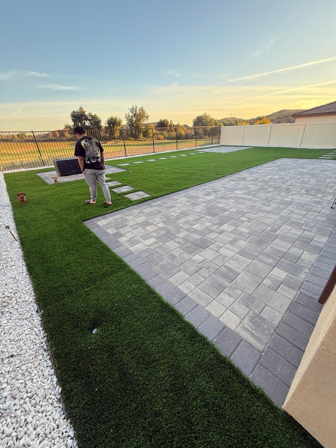 Gray paver patio bordered by artificial turf, with a white stone strip along the left side and a stepping path beyond