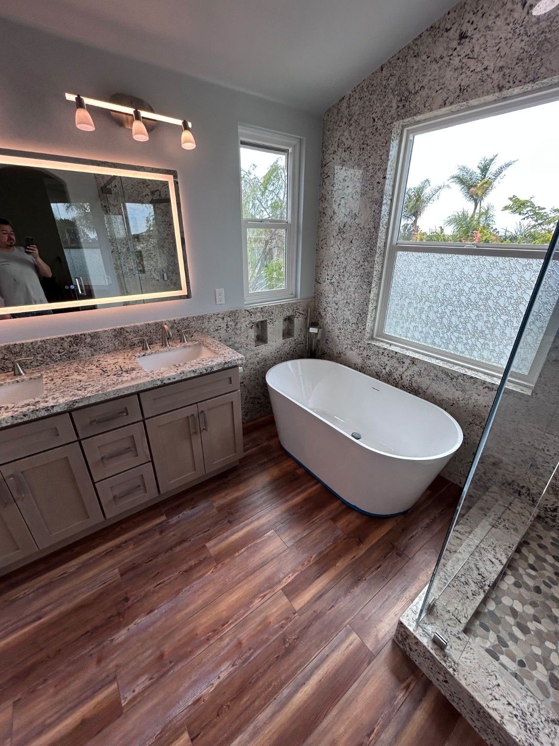 Double vanity with a lighted mirror next to a white freestanding tub beneath a window.
