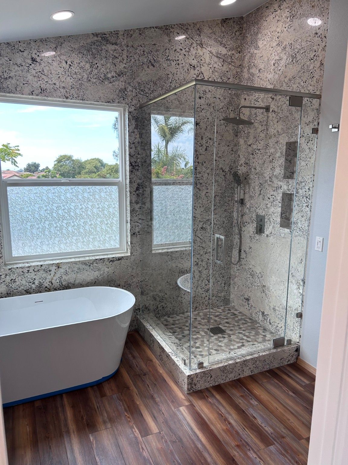 White freestanding tub beside a glass-enclosed shower with speckled walls, pebble-pattern floor and recessed niches.
