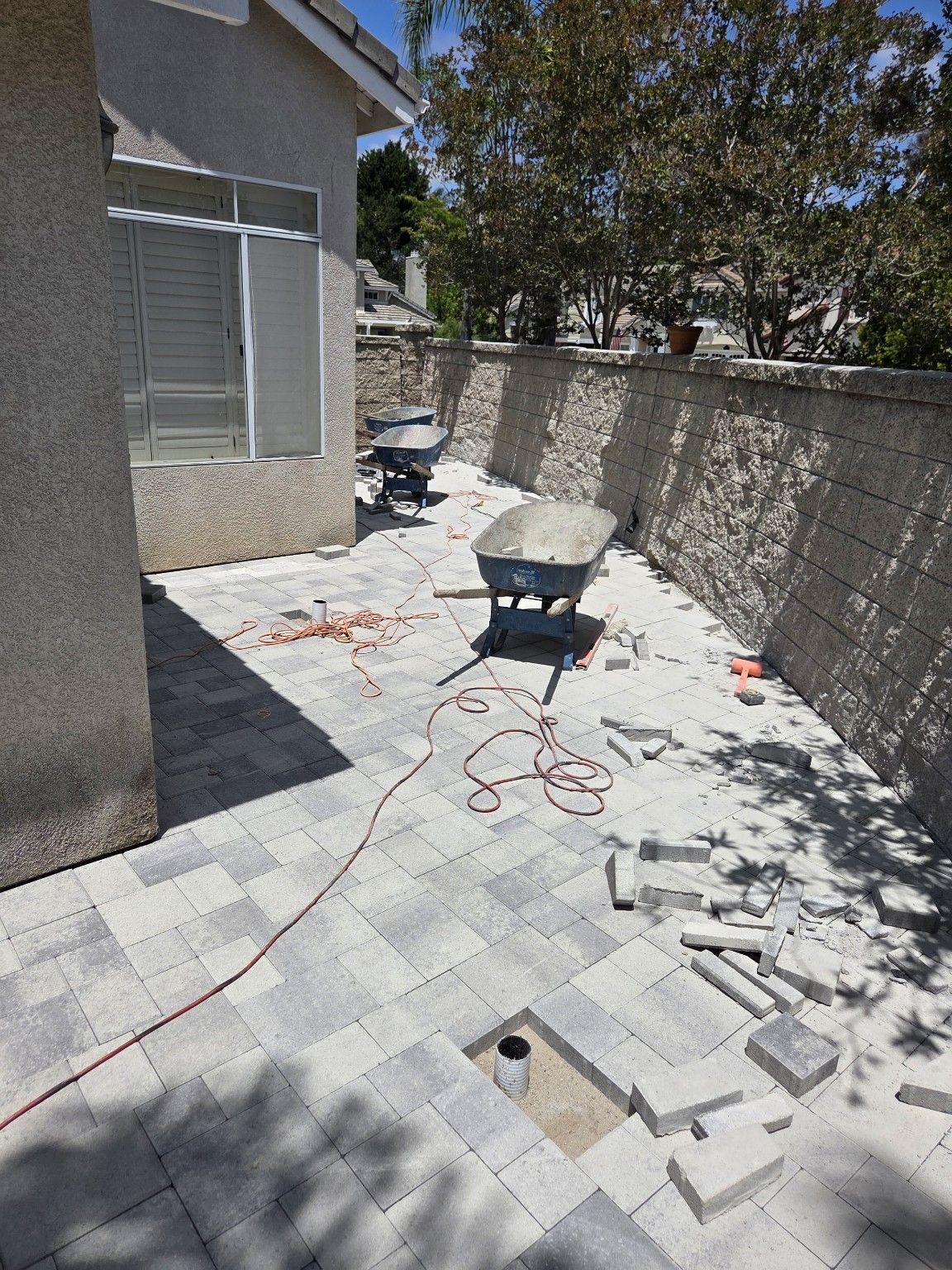 Side-yard pavers beside a block wall, with wheelbarrows, loose cut blocks and gaps around upright pipes