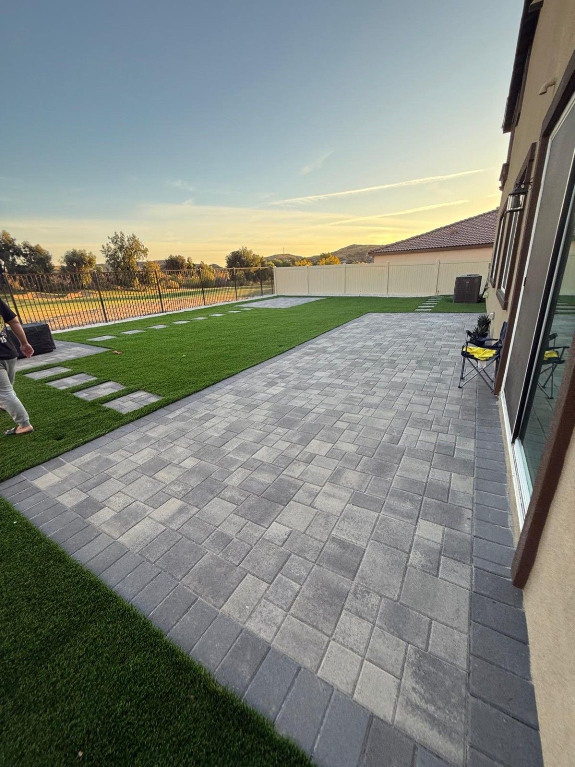 Long gray paver terrace beside the house’s sliding glass door, with artificial turf and separate paved areas beyond
