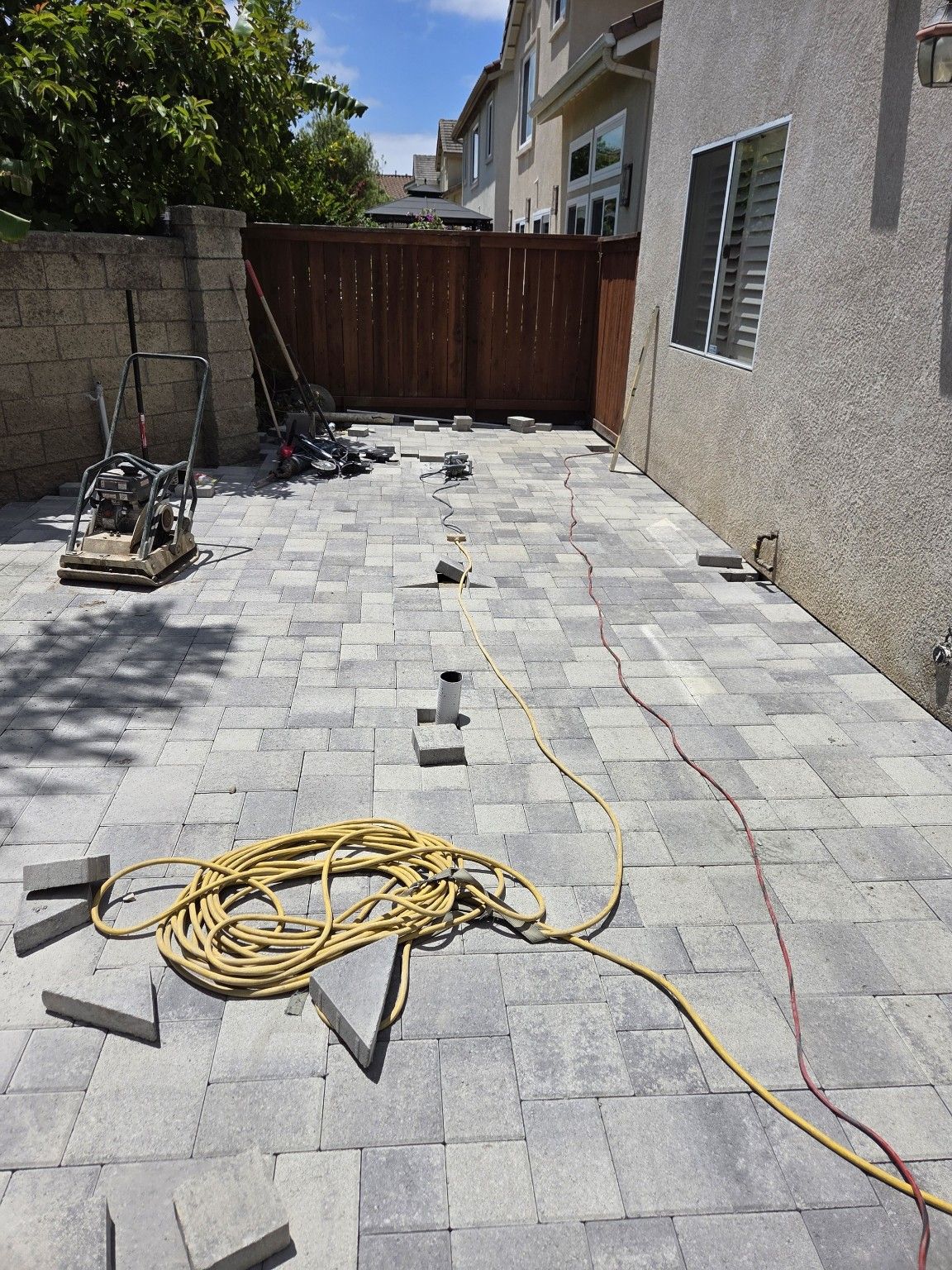 Gray pavers beside the house and wood gate, with a plate compactor, extension cords and loose paving blocks