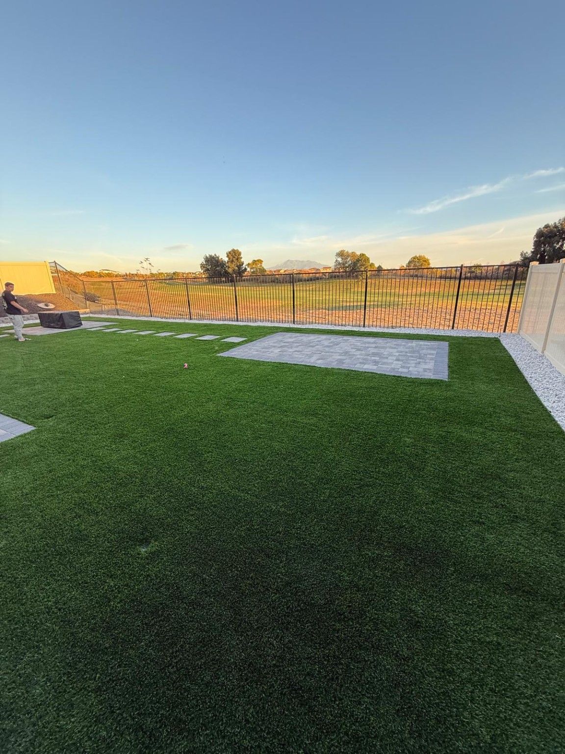 Rectangular gray paver pad set into artificial turf, connected by stepping stones beside a metal fence