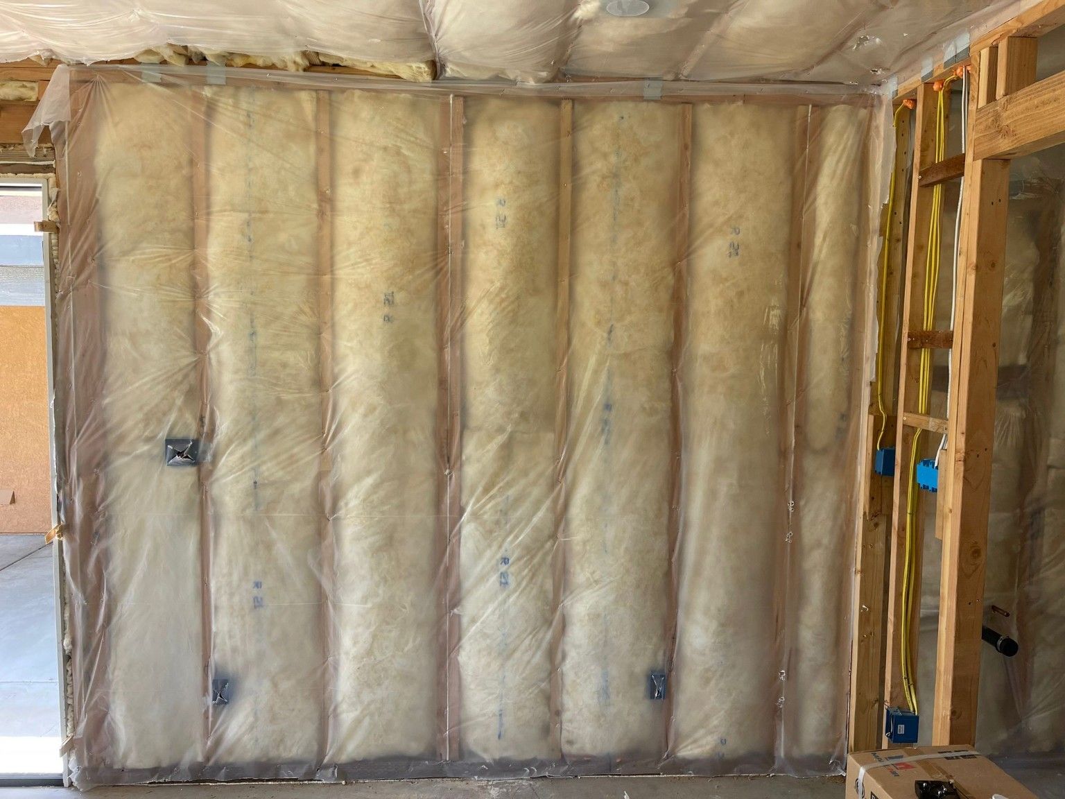 Insulation between wood wall studs beneath clear sheeting, with electrical wiring and boxes in exposed framing at right